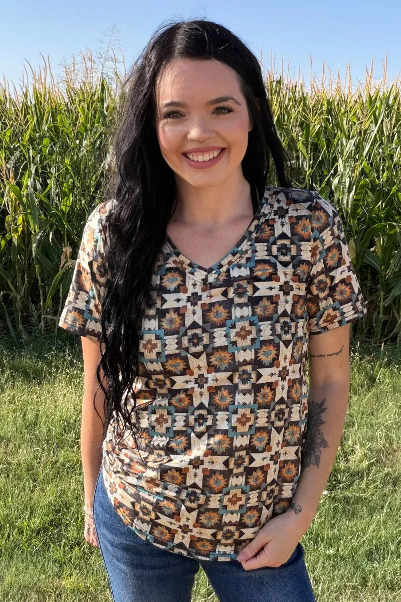 A woman with long dark hair stands outdoors in a grassy field with a cornstalks background, wearing a short-sleeve V-neck top with a geometric Southwestern-style pattern in beige, brown, blue, and black, paired with blue jeans.