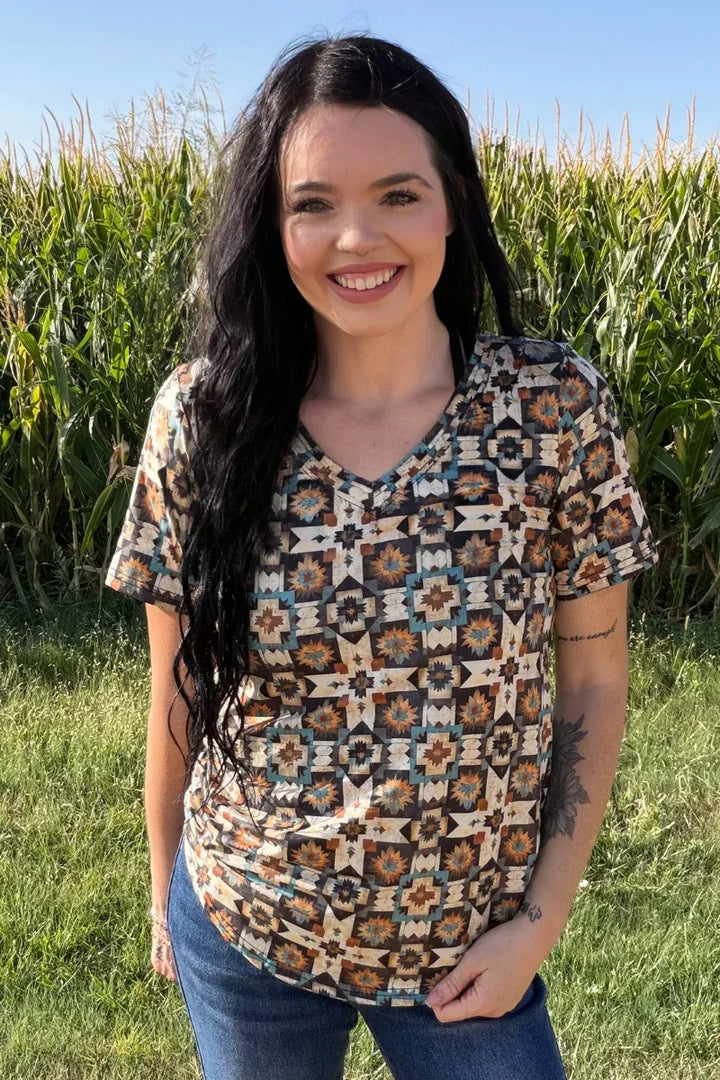 A woman with long dark hair stands outdoors in a grassy field with a cornstalks background, wearing a short-sleeve V-neck top with a geometric Southwestern-style pattern in beige, brown, blue, and black, paired with blue jeans.
