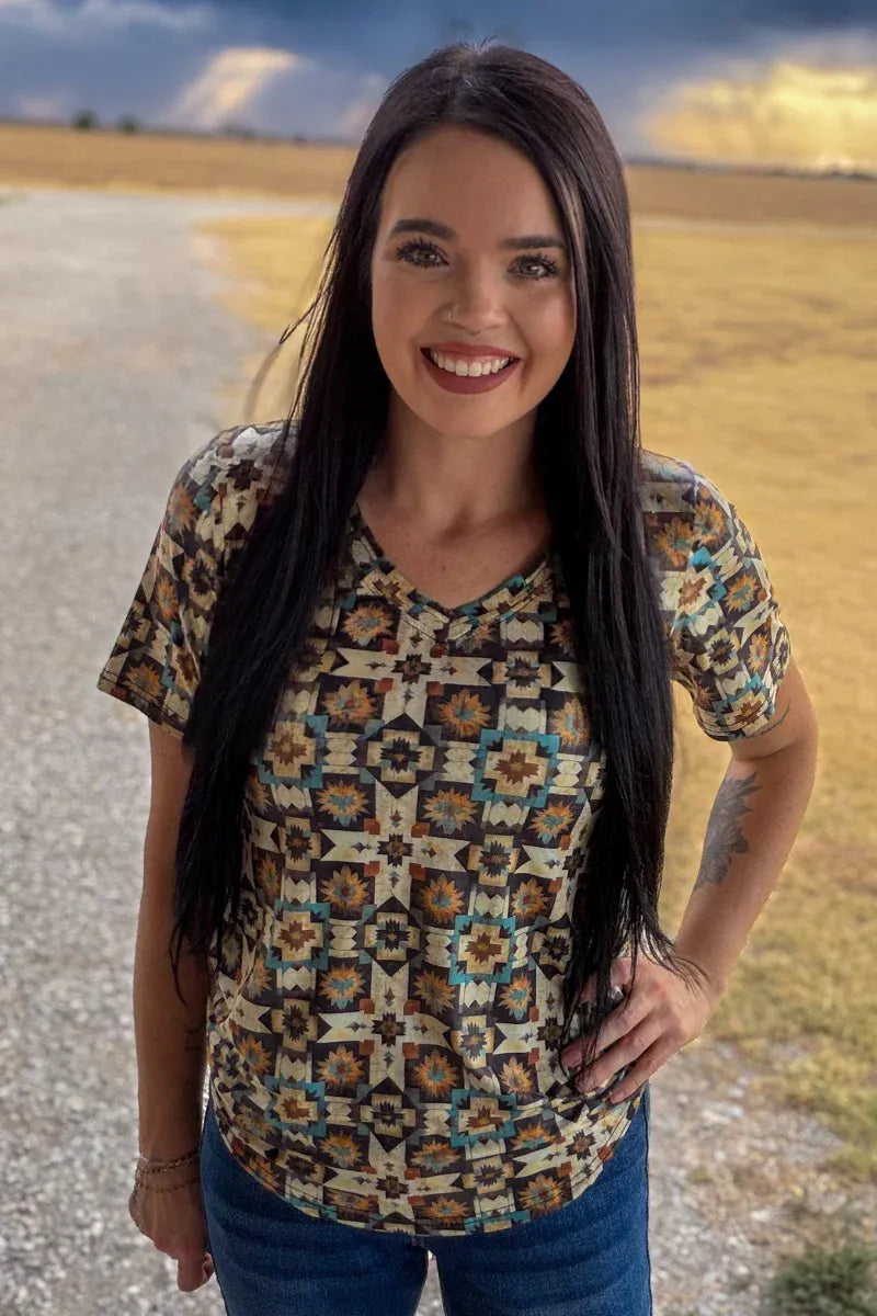A woman with long dark hair stands outdoors on a gravel path wearing a short-sleeve V-neck button-up shirt with a Southwestern geometric pattern in beige, brown, and teal, paired with blue jeans.