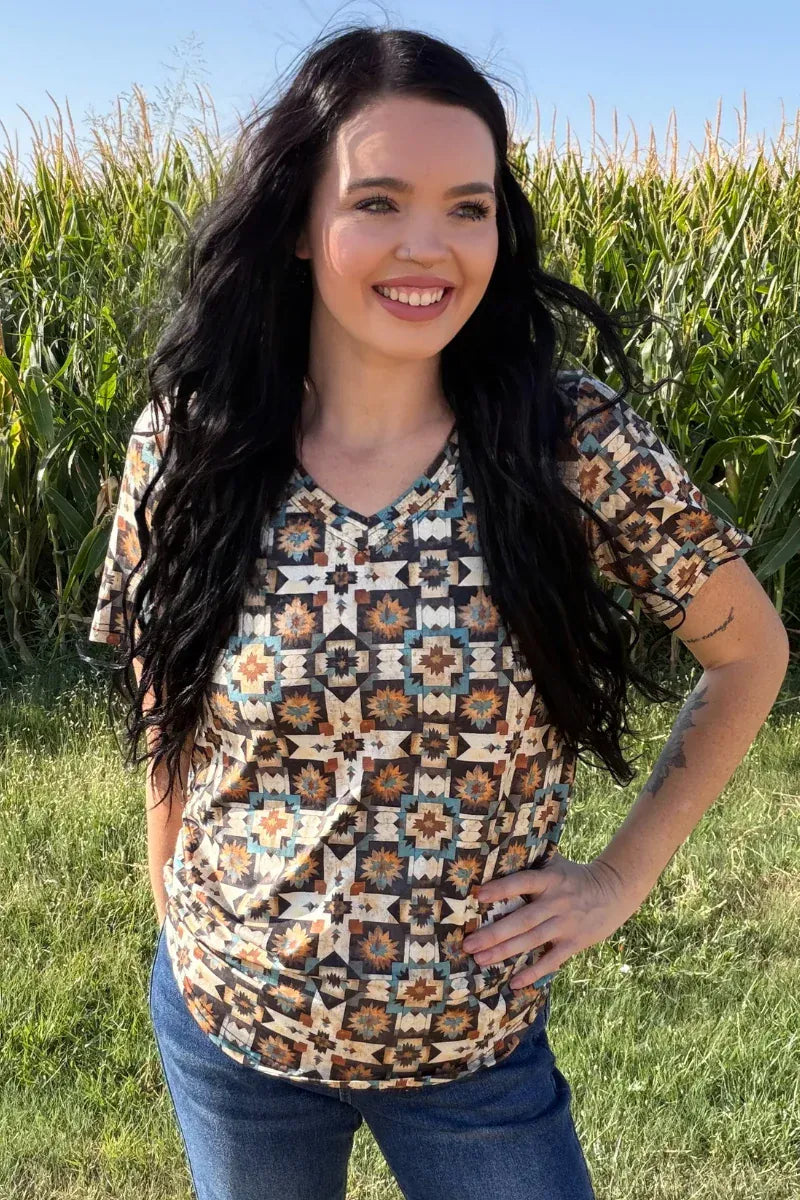 A woman with long dark hair stands outdoors in a grassy field with a cornstalks background, wearing a short-sleeve V-neck top with a geometric Southwestern-style pattern in brown, beige, and blue, paired with blue jeans.