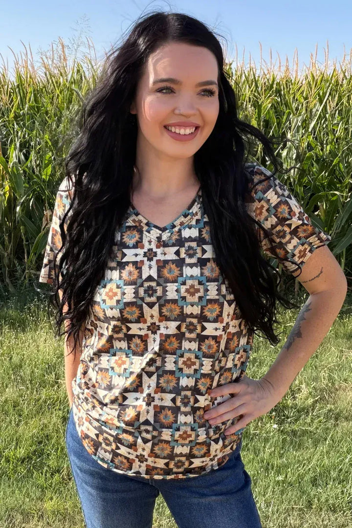 A woman with long dark hair stands outdoors in a grassy field with a cornstalks background, wearing a short-sleeve V-neck top with a geometric Southwestern-style pattern in brown, beige, and blue, paired with blue jeans.