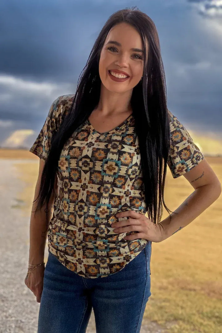 A woman with long dark hair stands outdoors wearing a short-sleeve V-neck top with a colorful geometric pattern in brown, beige, and blue, paired with blue jeans.