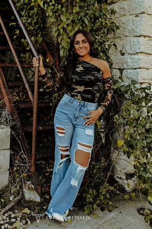 Woman wearing a camouflage top and ripped jeans standing against a stone wall with greenery.